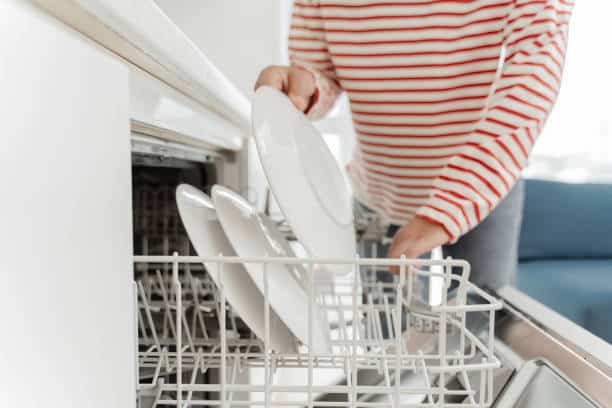 A person in a red and white striped shirt loads white plates into the bottom rack of an open dishwasher, showcasing the modern amenities at Delmont Townes in a bright kitchen.