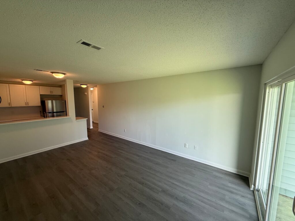 A spacious, unfurnished living area at Delmont Townes features wood-style flooring, white walls, a kitchen with appliances on the left, and sliding glass doors on the right—enjoy impressive amenities in every detail.