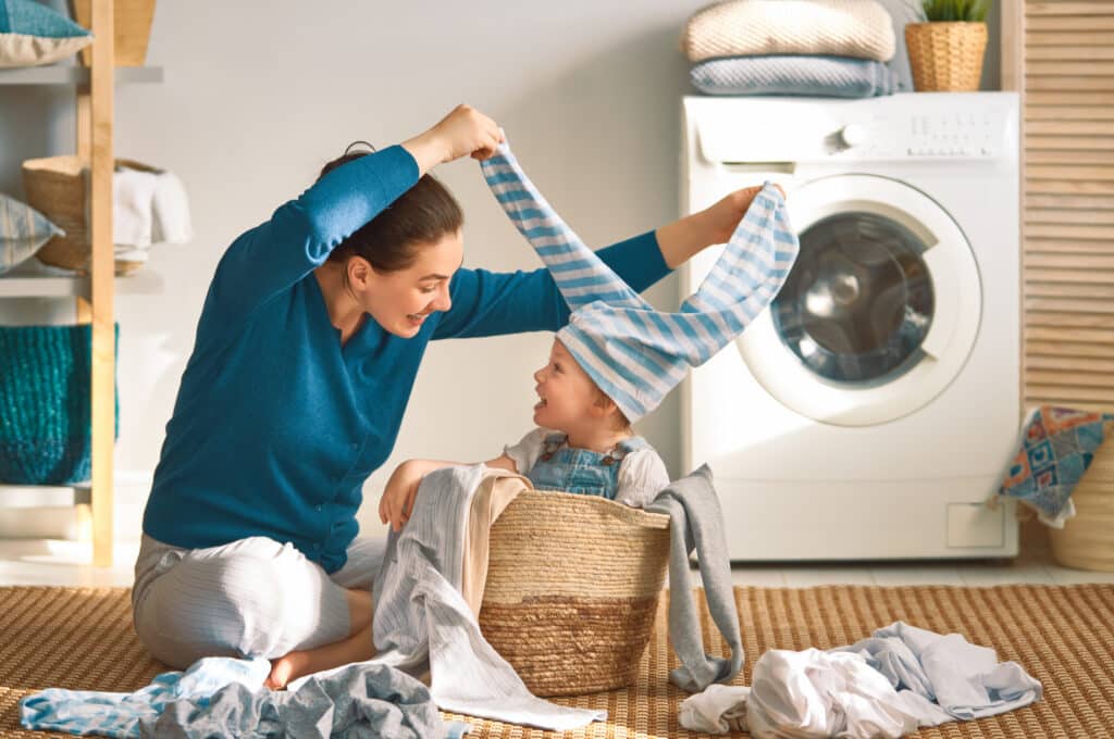 A woman and a child sit on the floor next to a laundry basket and washing machine at Delmont Townes, enjoying quality time together as they play with a striped shirt—one of many family-friendly amenities.