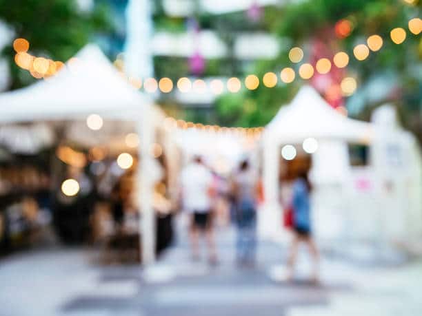 Blurred outdoor market scene with white tents, string lights overhead, and people walking among stalls—capturing the vibrant community atmosphere and local amenities near Delmont Townes.