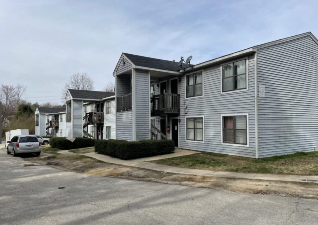 Gallery - Delmont Townes features two-story gray apartment buildings with exterior staircases, small balconies, and a parked car on a paved lot under a cloudy sky.