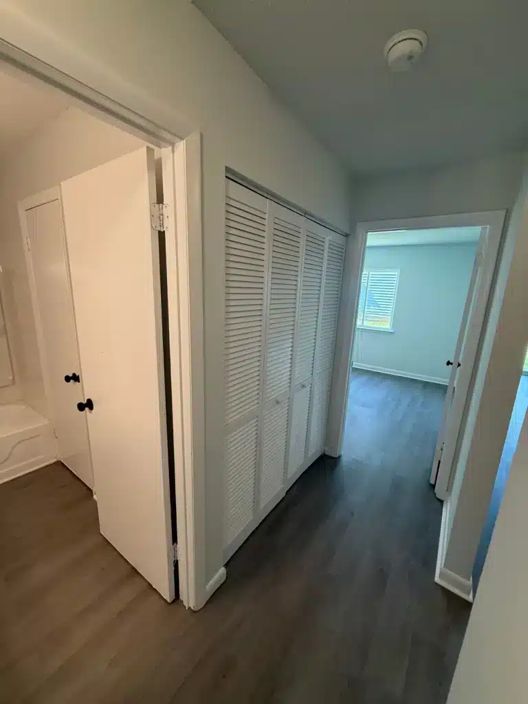 A hallway with wood-style flooring, white doors to a bathroom and closets on the left, and open doorways leading to two rooms with windows ahead in Gallery - Delmont Townes.