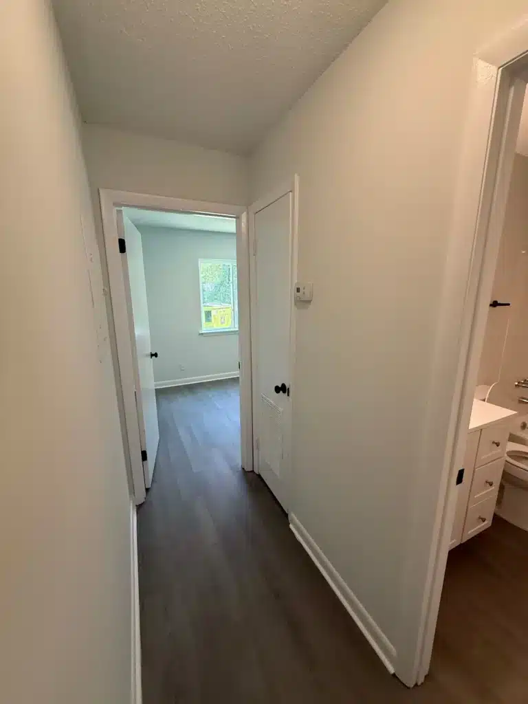 A narrow hallway with white walls and dark wood flooring at Gallery - Delmont Townes leads to a bedroom with a window; a bathroom with a toilet and sink is visible on the right.