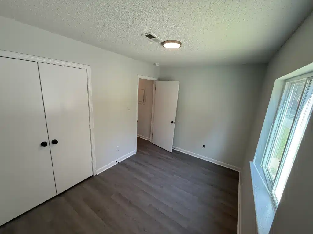 Empty room with light gray walls, white closet doors, wood-look flooring, a ceiling light, window, and an open door leading to another space—perfect for showcasing your style at Gallery - Delmont Townes.