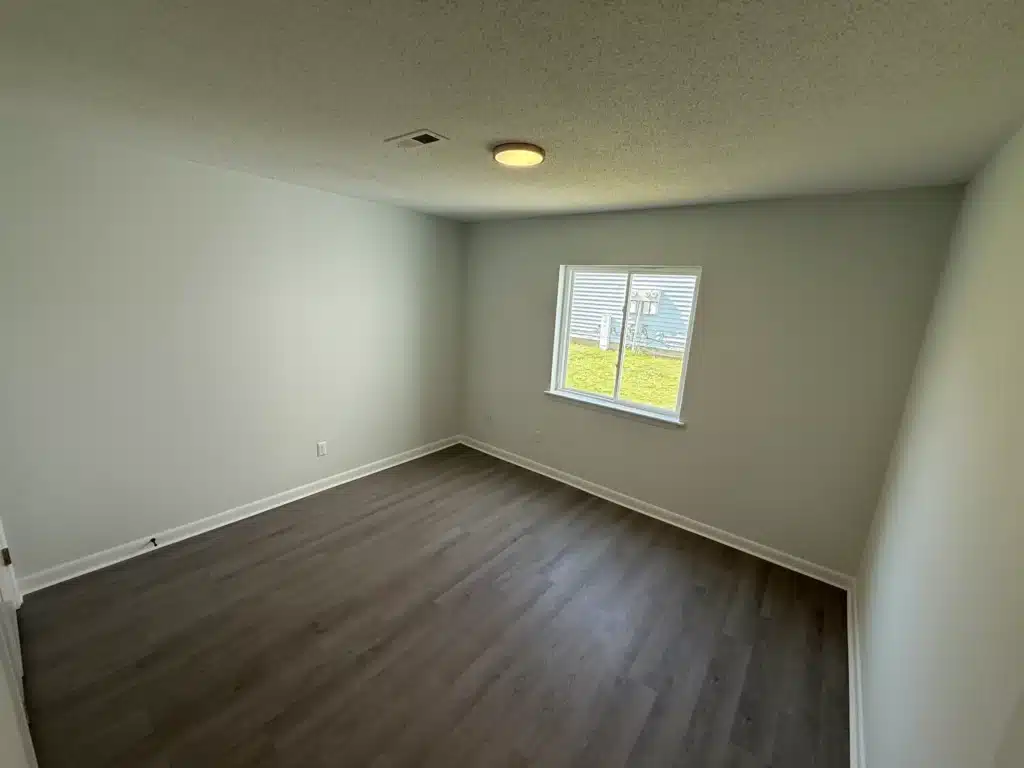 Empty room at Gallery - Delmont Townes with light gray walls, dark wood flooring, a ceiling light, and a window letting in natural light, overlooking a grassy area outside.