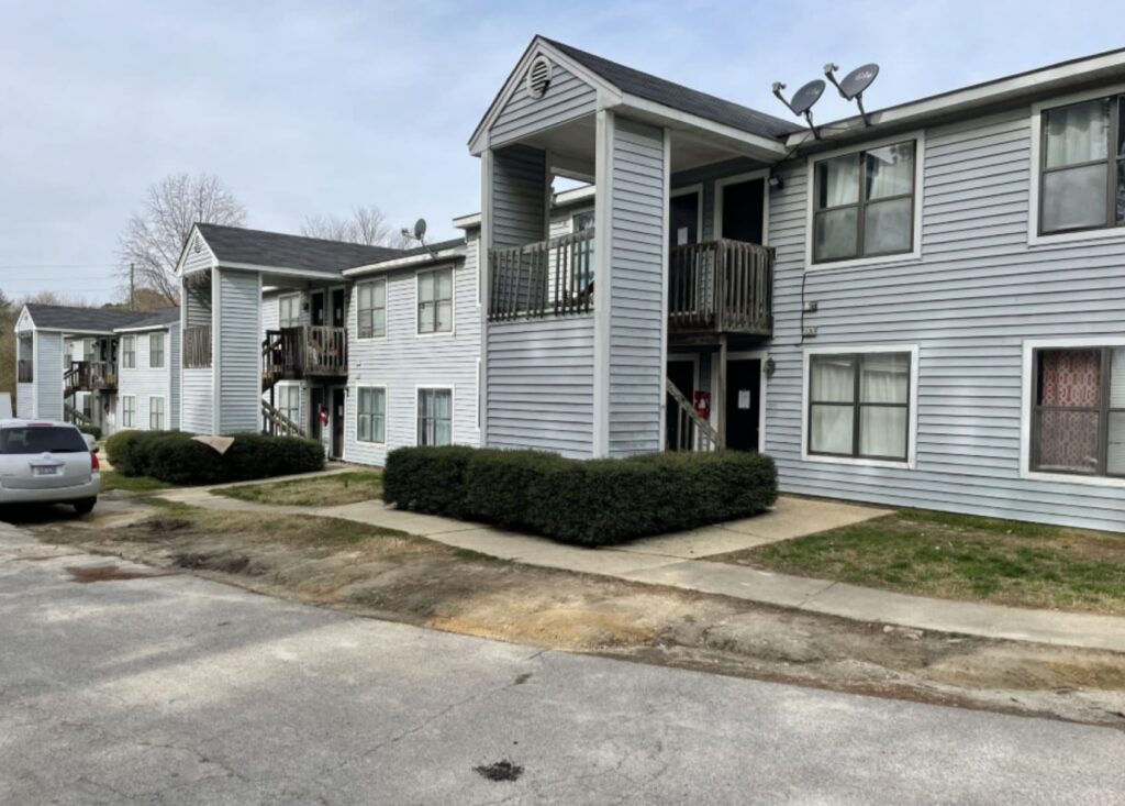 Gallery - Delmont Townes features a two-story apartment building with gray siding, small balconies, and satellite dishes on the roof, all viewed from a convenient parking lot.