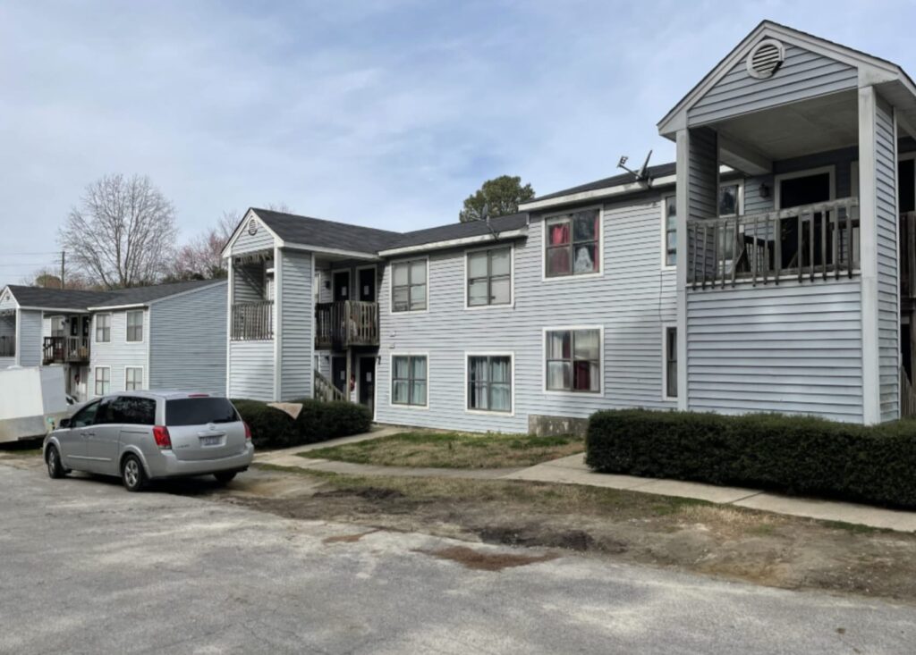 Gallery - Delmont Townes features a two-story gray apartment building with balconies and a parked silver minivan in front on a partly cloudy day.
