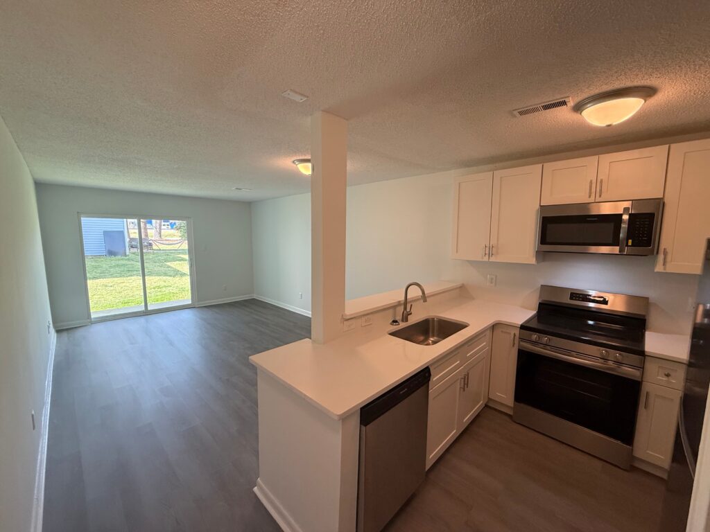 Modern kitchen with stainless steel appliances opens to a spacious living area in Gallery - Delmont Townes, with sliding glass doors leading to a grassy backyard.