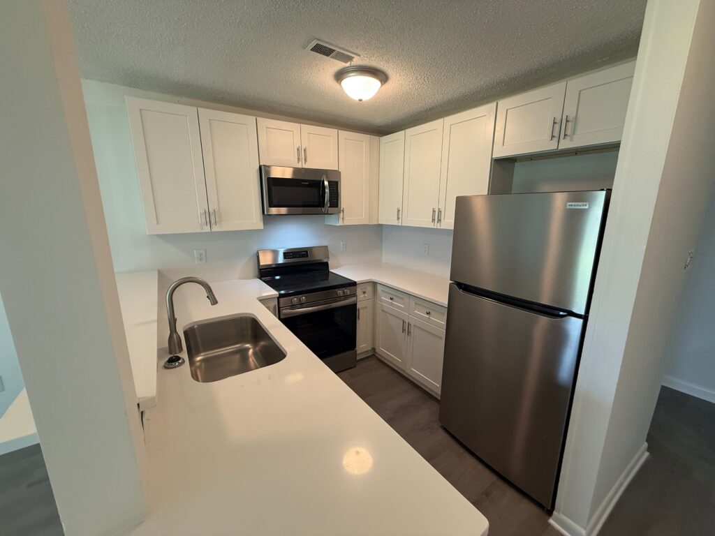 Modern kitchen at Gallery - Delmont Townes with white cabinets, stainless steel appliances, electric stove, built-in microwave, double sink, and sleek white countertops under a bright ceiling light.