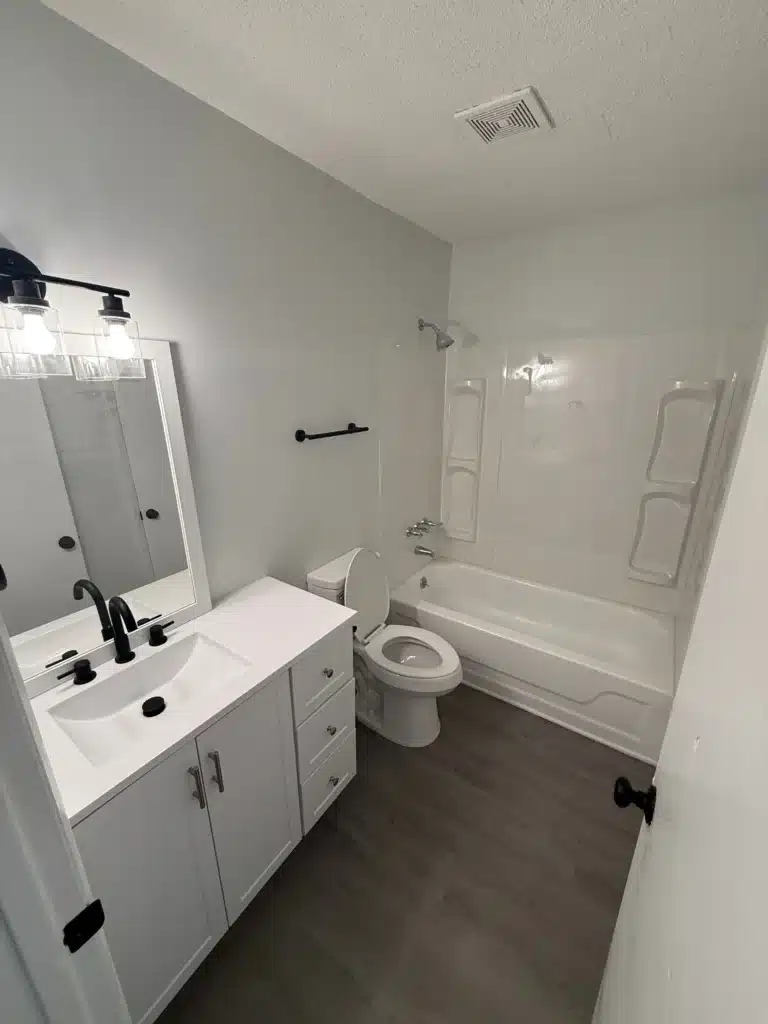 A modern bathroom at Gallery - Delmont Townes features a white vanity, sink, toilet, and a bathtub-shower combination. The floor has gray wood-style flooring and the walls are painted light gray for a sleek, contemporary look.